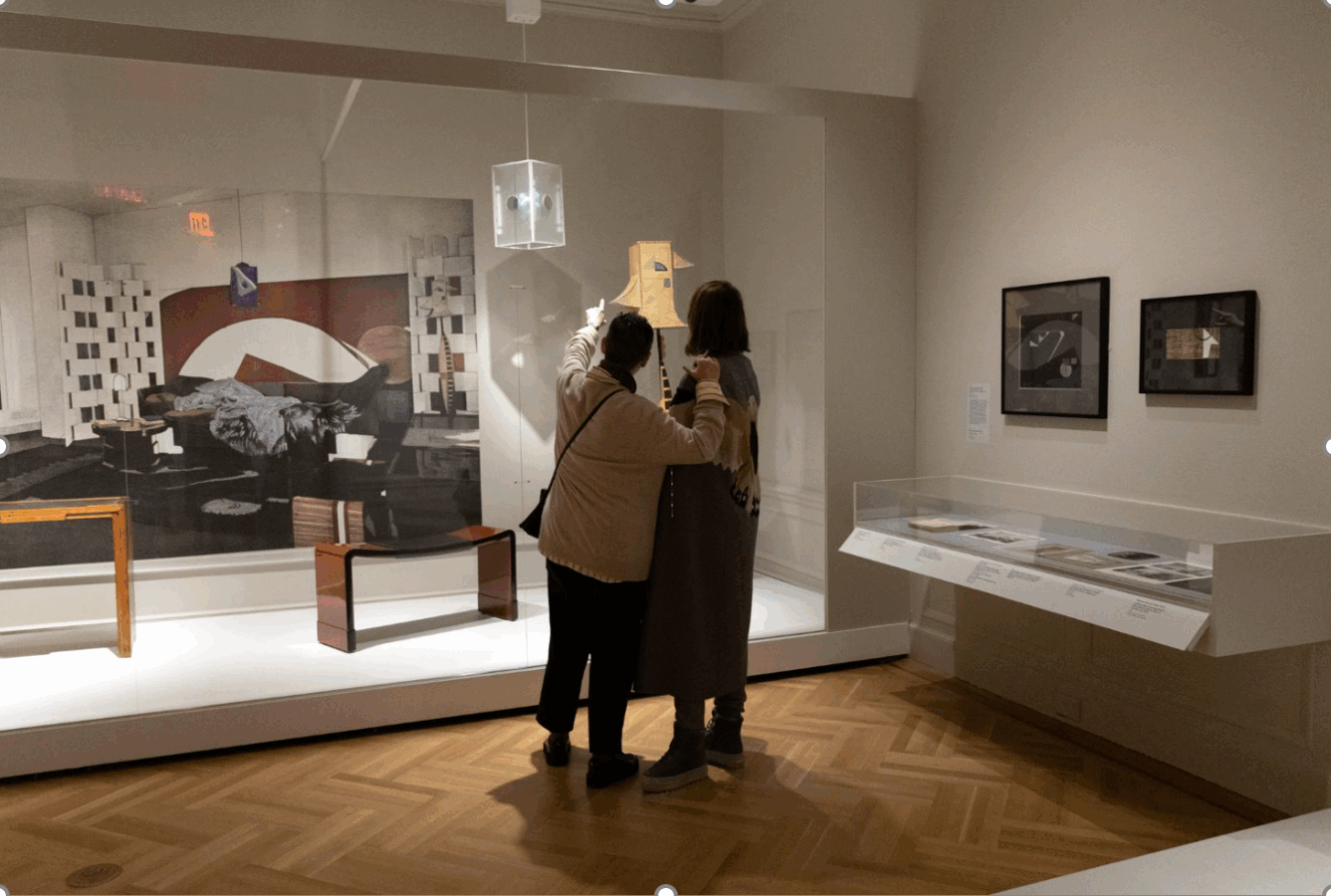 Two people standing close together in an art museum, pointing toward a sculptural object on display within a modernist exhibit space.
